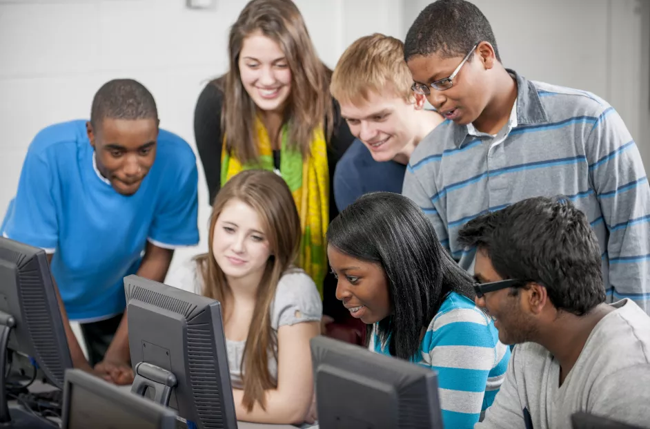 A multi-ethnic group of high school age students are gathered around a computer and are watching a video together in the lab.