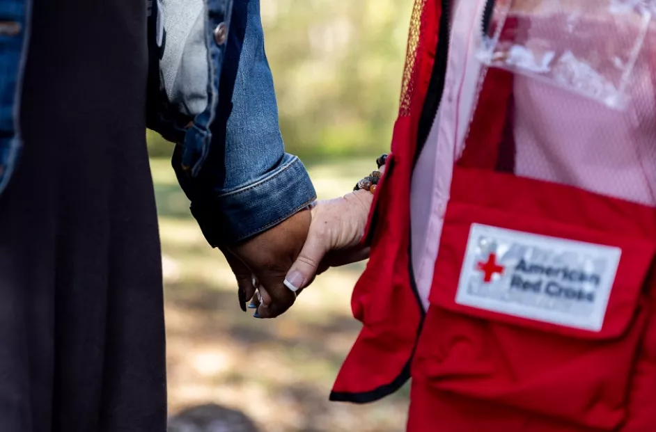 Close-up image of two people holding hands. One person is wearing a denim jacket, while the other is wearing a red vest with an American Red Cross patch. The background is blurred, suggesting an outdoor setting.