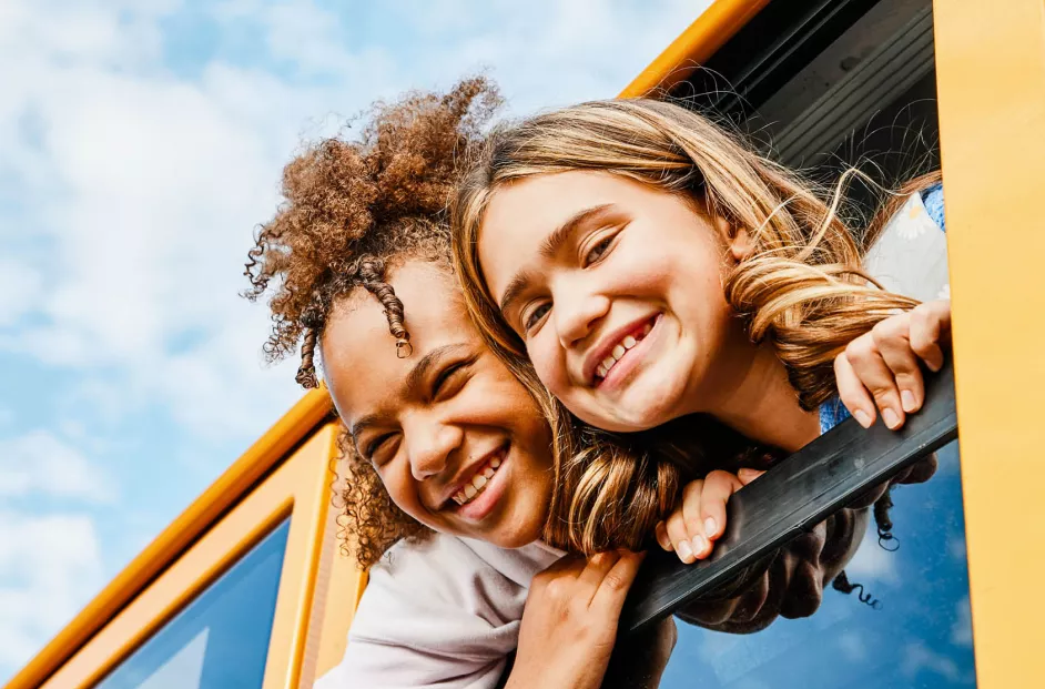 Two girls smile as they hang their heads out of the window of a bus.