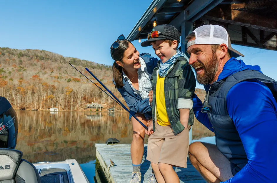 Three people stand on a wooden dock beside a calm lake, with one adult helping a child step toward a small boat while another adult steadies them. Fishing rods are visible, and wooded hills with autumn foliage reflect on the water in the background.