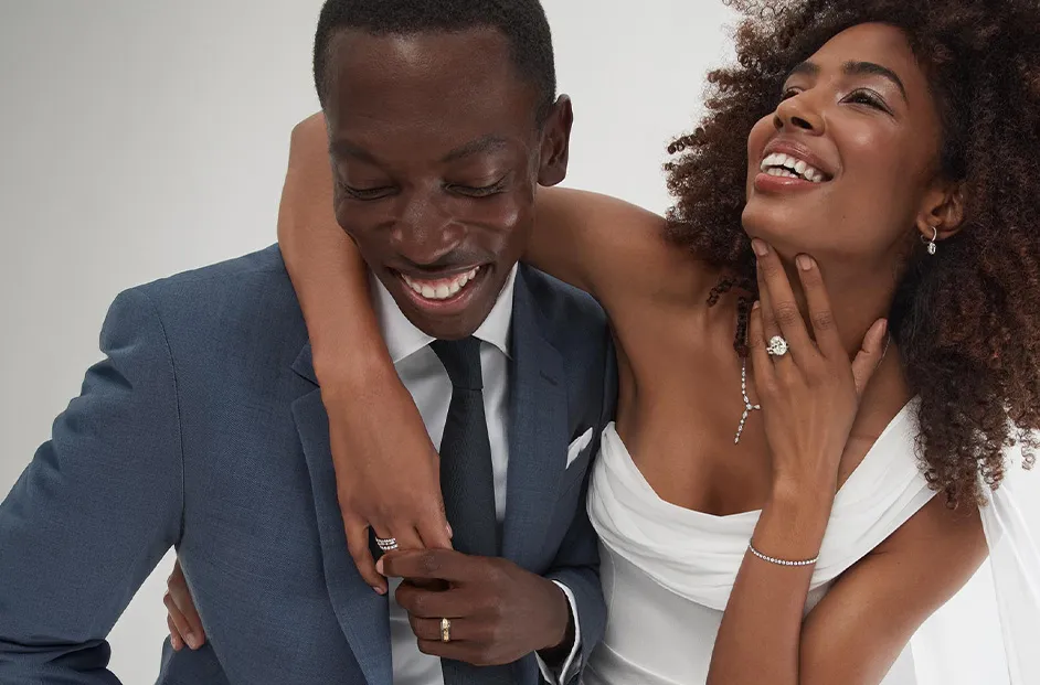 Two people in formal attire pose together while wearing wedding and fine jewelry, including engagement rings, wedding bands, bracelets, earrings, and necklaces against a light background.
