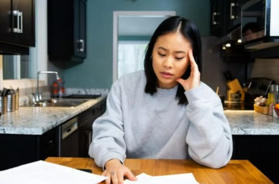 A young Asian woman looks stressed while working on paperwork at her kitchen table.