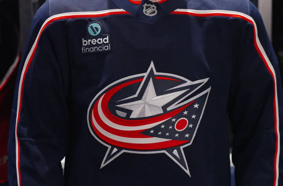 A Columbus Blue Jackets hockey jersey hangs in a locker inside a team dressing room. The navy blue jersey features the Blue Jackets crest on the front, the number 8 on both sleeves, an assistant captain "A" patch on the chest, and a Bread Financial logo patch positioned on the upper left side of the jersey. The locker area is dimly lit, drawing focus to the jersey and its details.