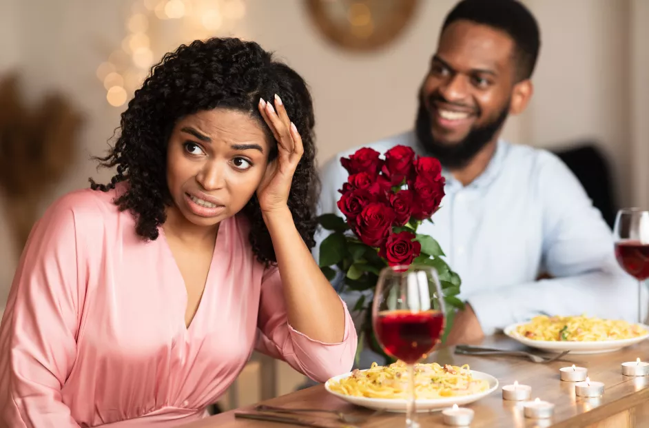 Image of a man and woman on a dinner date. The woman has a pained expression on her face and her back turned to the man, who is smiling. Plates of spaghetti and glasses of wine are untouched in front of the pair, along with a bouquet of roses.