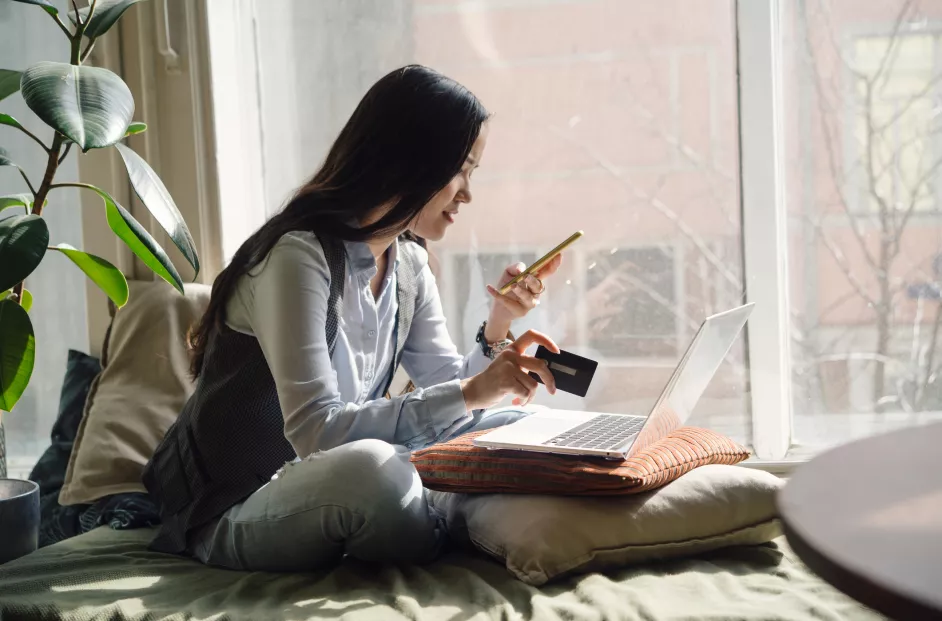 A young Asian woman in blue jeans sitting on the bed in a yoga pose in front of a laptop. Online shopping with her phone and credit card.