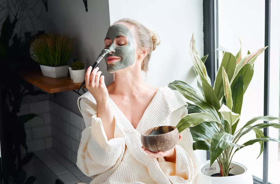 Positive young female in white bathrobe applying purifying clay mask on face with brush from bowl while sitting near window and relaxing during skincare procedure at home