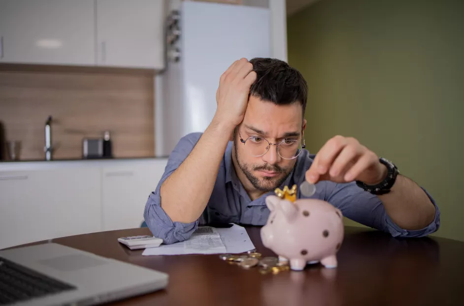Young man with glasses puts money into a piggy bank. He is stressed.