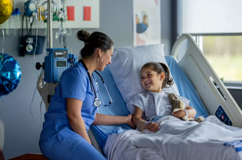 A female nurse makes her rounds to check in on patients after their surgery. She is sitting with a sweet little girl who is recovering in her hospital bed and cuddling her stuffed animal as she talks with her.