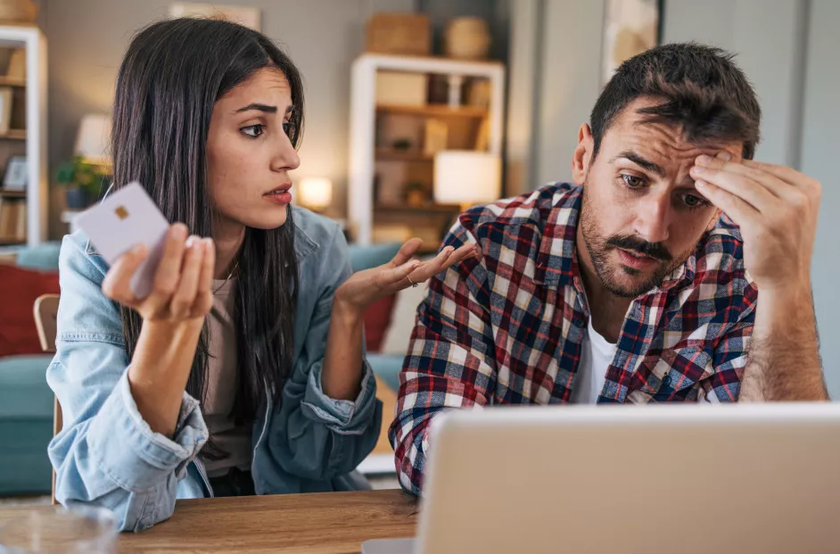 An adult cheerful couple sitting at the table, using a laptop, with the living room visible in the background. They are shopping online with a credit card and look concerned.