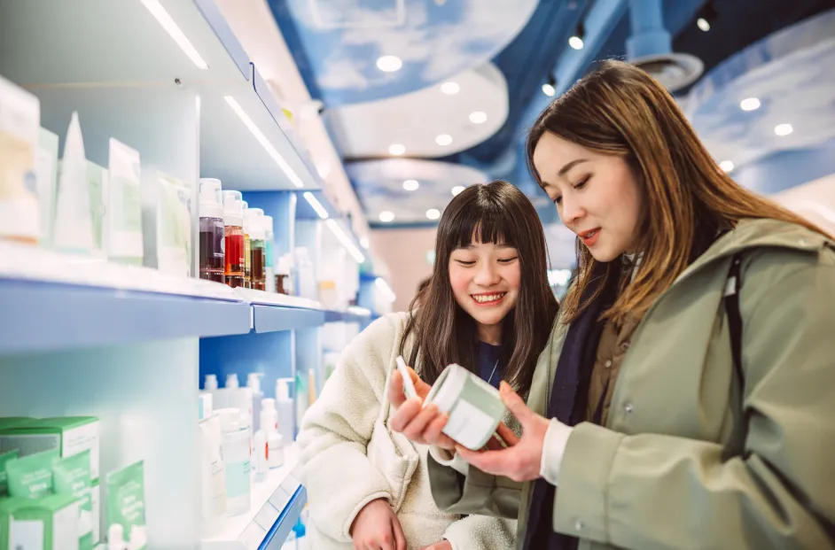 Smiling mother and daughter browsing skincare products together in a modern beauty store. The teenage daughter looks on with curiosity as her mother carefully reads a product label, highlighting family bonding, everyday wellness, and conscious consumer choices. Ideal for concepts related to skincare, family life, sustainability, and mindful shopping.