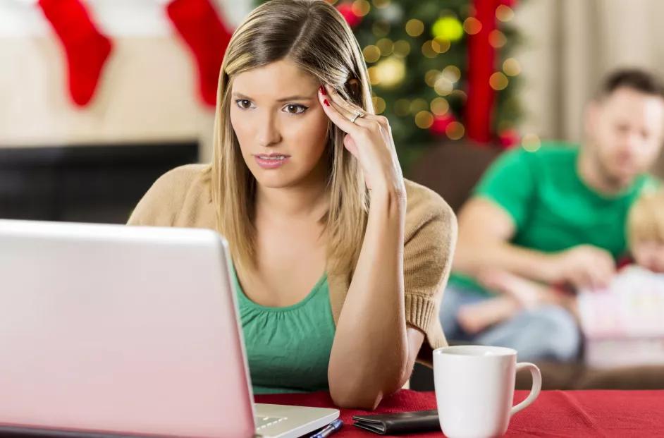 Woman appears stressed while using her laptop. Holiday tree and stockings in the background.
