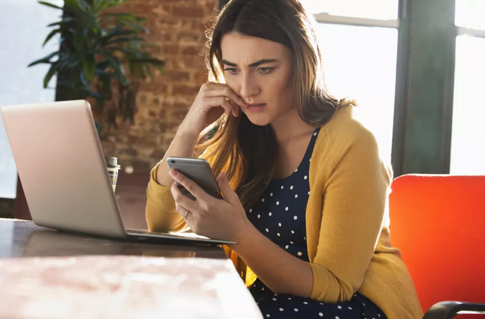 A woman looks concerned while doing work on her computer and phone.