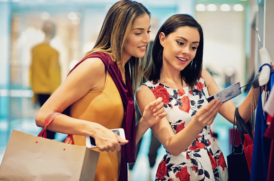 Two people browse clothing on a rack inside a retail store, holding shopping bags and a smartphone while looking at a garment tag in a bright, modern shopping environment.