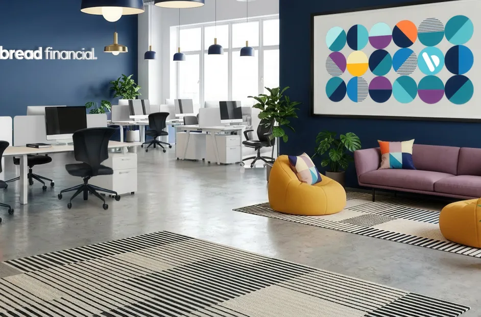 A modern Bread Financial office featuring an open workspace with rows of desks, computer monitors, and ergonomic chairs. The space has navy blue accent walls, large windows, pendant lighting, and several green plants. A lounge area in the foreground includes a purple sofa, yellow lounge chairs, patterned rugs, and colorful geometric accent pillows. The Bread Financial logo is displayed on the wall, alongside framed artwork featuring the company's circular brand design elements.