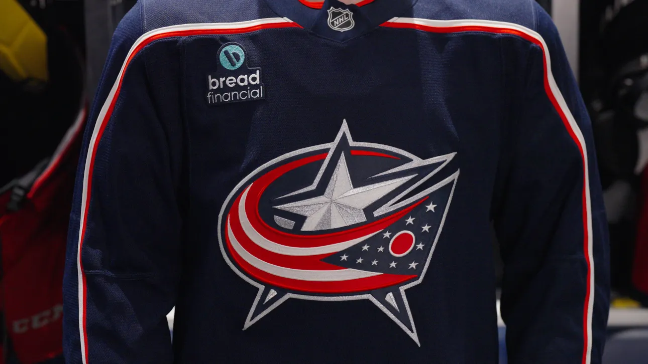 A Columbus Blue Jackets hockey jersey hangs in a locker inside a team dressing room. The navy blue jersey features the Blue Jackets crest on the front, the number 8 on both sleeves, an assistant captain "A" patch on the chest, and a Bread Financial logo patch positioned on the upper left side of the jersey. The locker area is dimly lit, drawing focus to the jersey and its details.