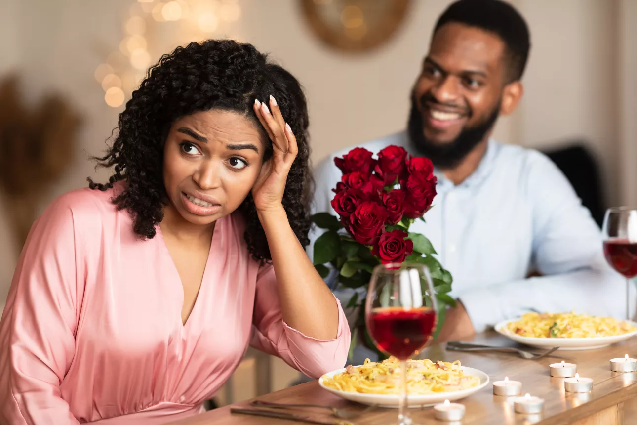 Image of a man and woman on a dinner date. The woman has a pained expression on her face and her back turned to the man, who is smiling. Plates of spaghetti and glasses of wine are untouched in front of the pair, along with a bouquet of roses.