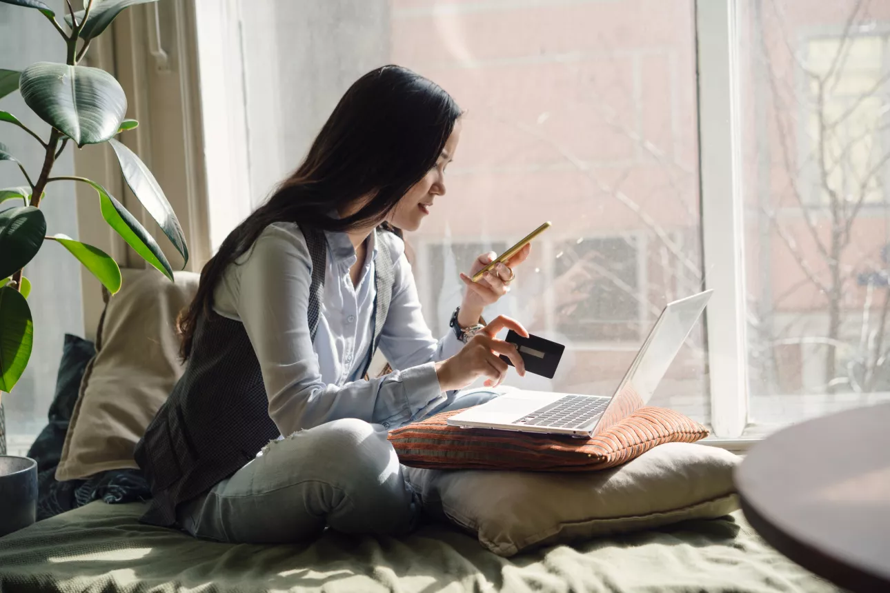 A young Asian woman in blue jeans sitting on the bed in a yoga pose in front of a laptop. Online shopping with her phone and credit card.