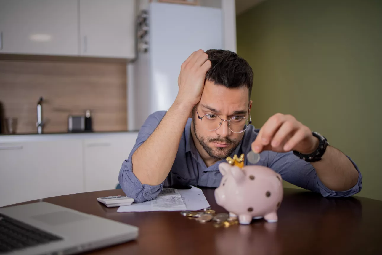 Young man with glasses puts money into a piggy bank. He is stressed.