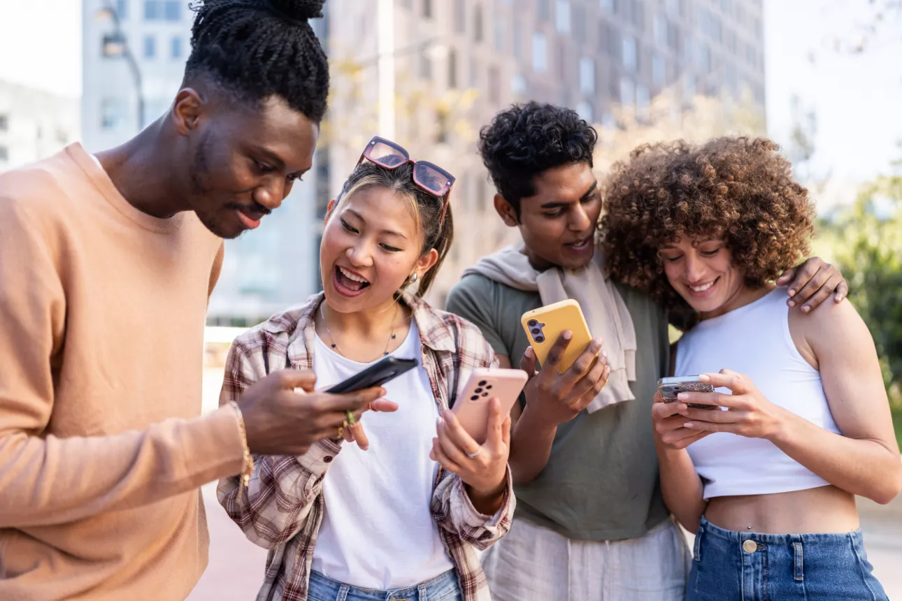 Four friends are gathered together outdoors, each engrossed in their own smartphones. They seem to be sharing content with each other, smiling and interacting as they stand on a city street with buildings in the background on a bright, sunny day.