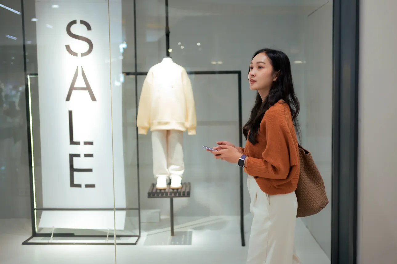 Young woman holding smartphone, pausing to browse a boutique window display with Sale sign, contemplating purchases in mall