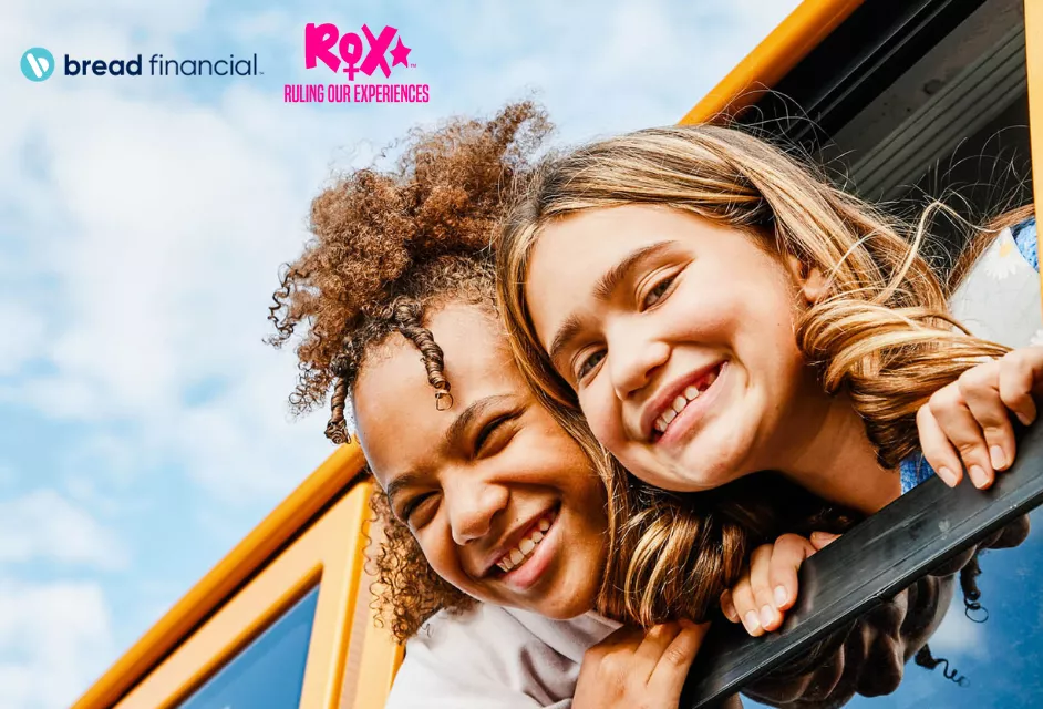 Two girls smile as they hang their heads out of the window of a bus.